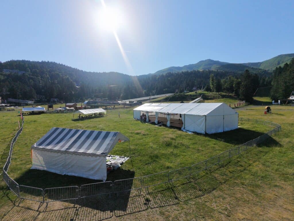 Tentes d'événement blanches sur herbe en montagne sous un soleil radieux. Clôtures métalliques au premier plan.