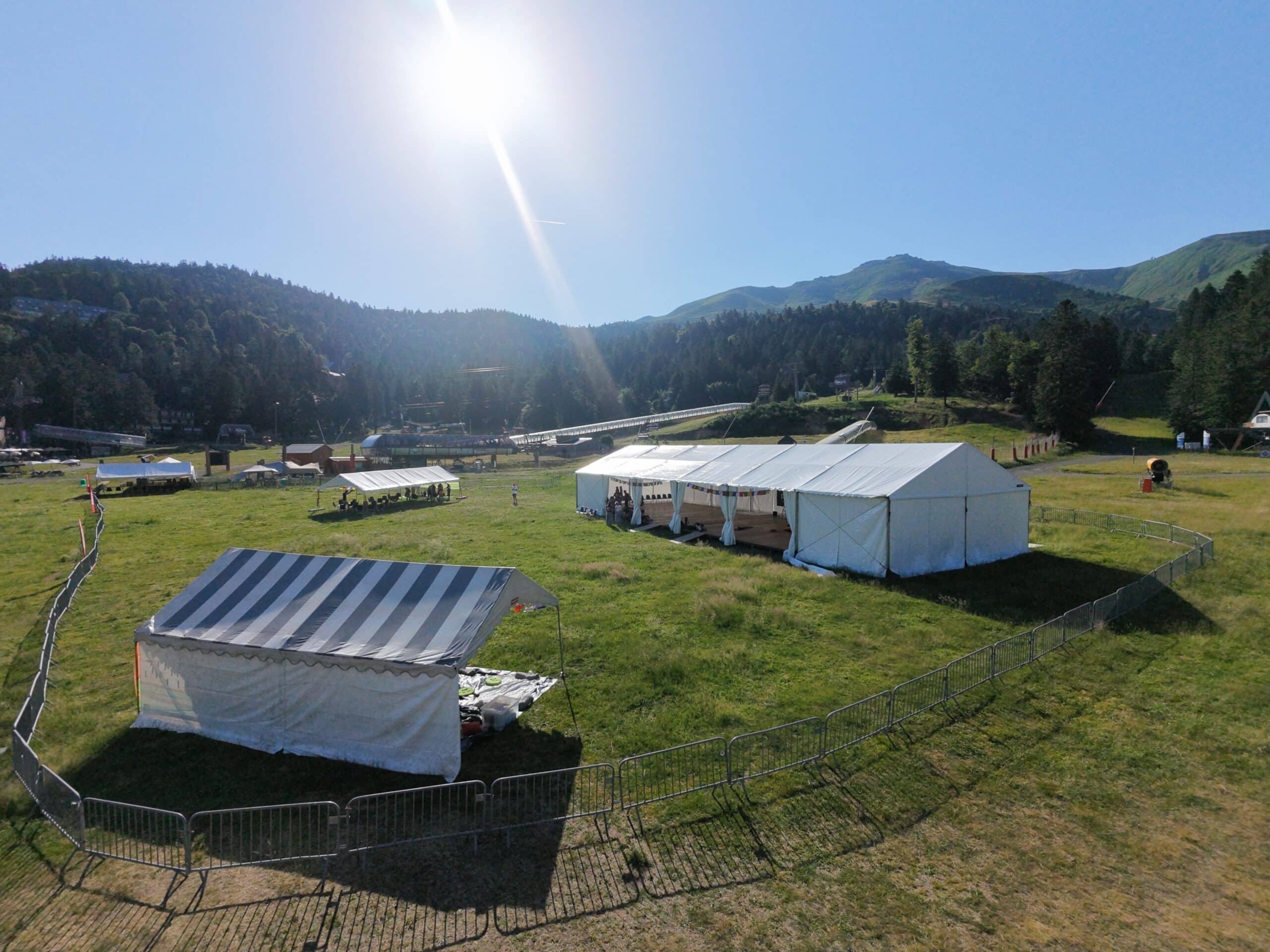 Tentes d'événement blanches sur herbe en montagne sous un soleil radieux. Clôtures métalliques au premier plan.