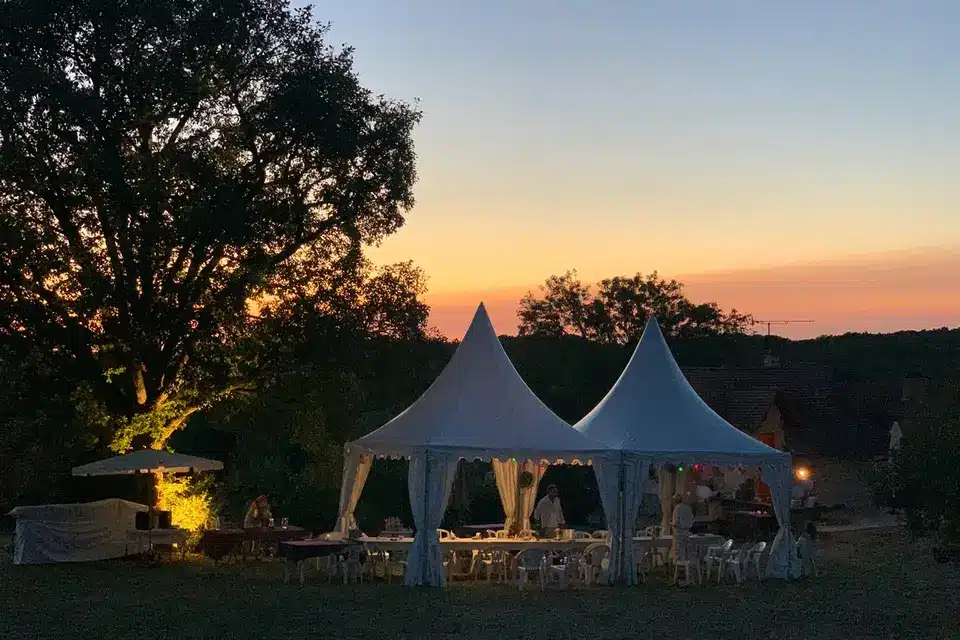 Tentes événementielles blanches sous un ciel de coucher de soleil orangé, avec un grand arbre illuminé à gauche, ambiance de fête en plein air.