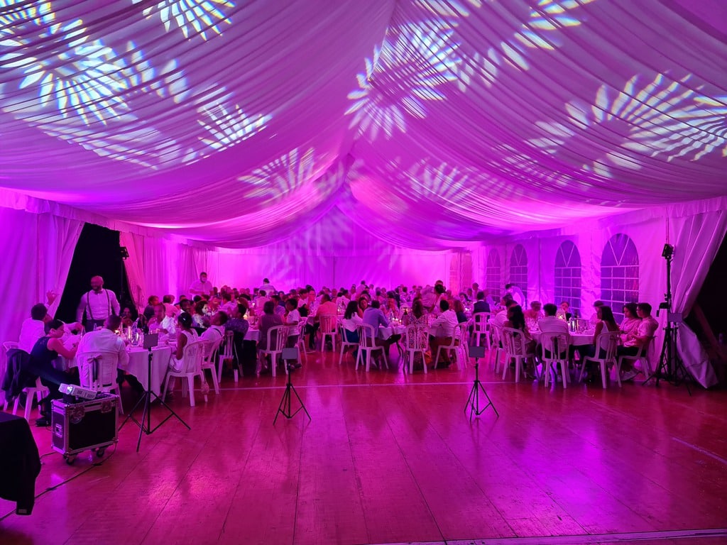 Fête sous chapiteau éclairé en violet, avec projections de motifs végétaux sur la toile, invités assis à table pour un dîner.