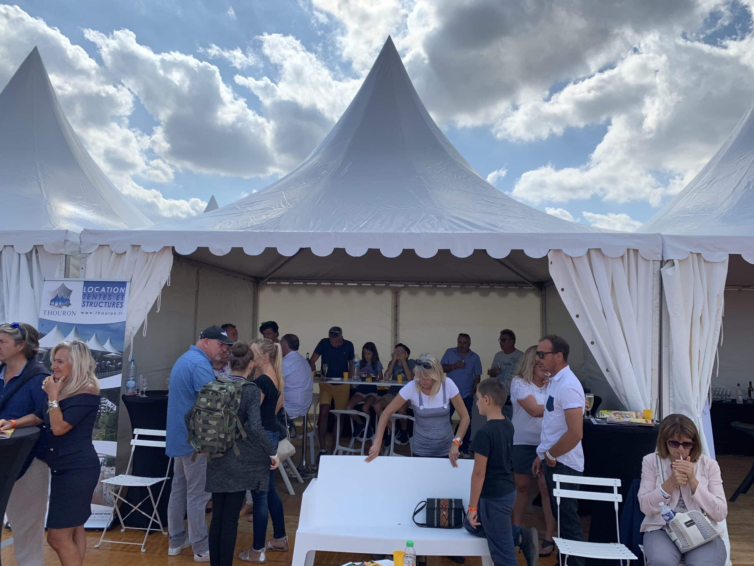Fête sous une grande tente blanche au soleil, foule discutant près d'un panneau "LOCATION TENTES ET STRUCTURES THOURON".