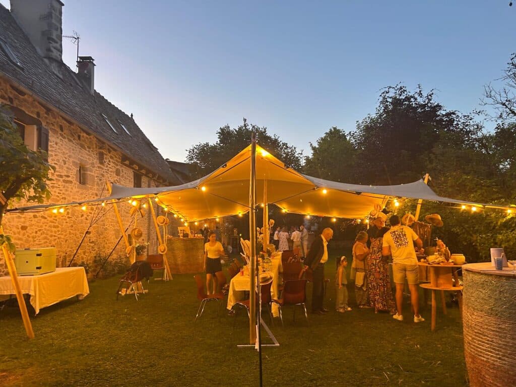 Réception festive sous une tente stretch éclairée par des guirlandes dans le jardin d'une maison en pierre.
