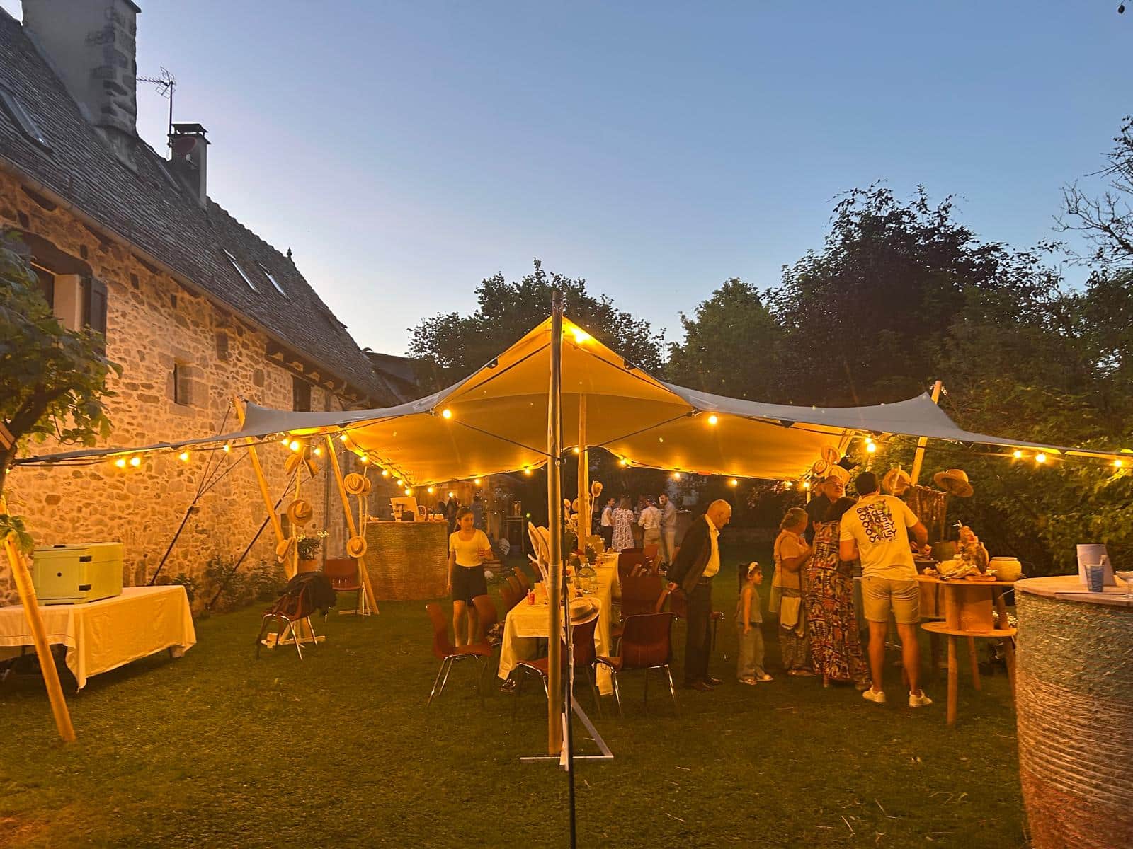 Réception festive sous une tente stretch éclairée par des guirlandes dans le jardin d'une maison en pierre.
