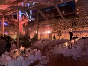 Tente transparente élégante pour dîner de réception avec tables dressées, chaises cristal, arbre illuminé et mur de pierres apparentes.