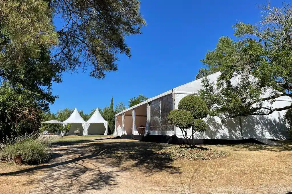 Grande tente blanche événementielle et petits chapiteaux blancs dans un cadre extérieur ensoleillé, entourés d'arbres et de végétation.