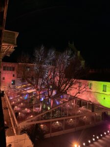 Tente transparente de réception illuminée dans une cour de château la nuit, avec un arbre central et des murs colorés en rouge et vert.