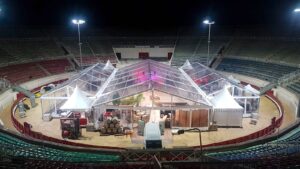 Installation d'une grande tente transparente dans une arène de nuit, vue d'en haut, avec futs de vin et chariots élévateurs. Texte visible: VILLE DE BEZIERS.