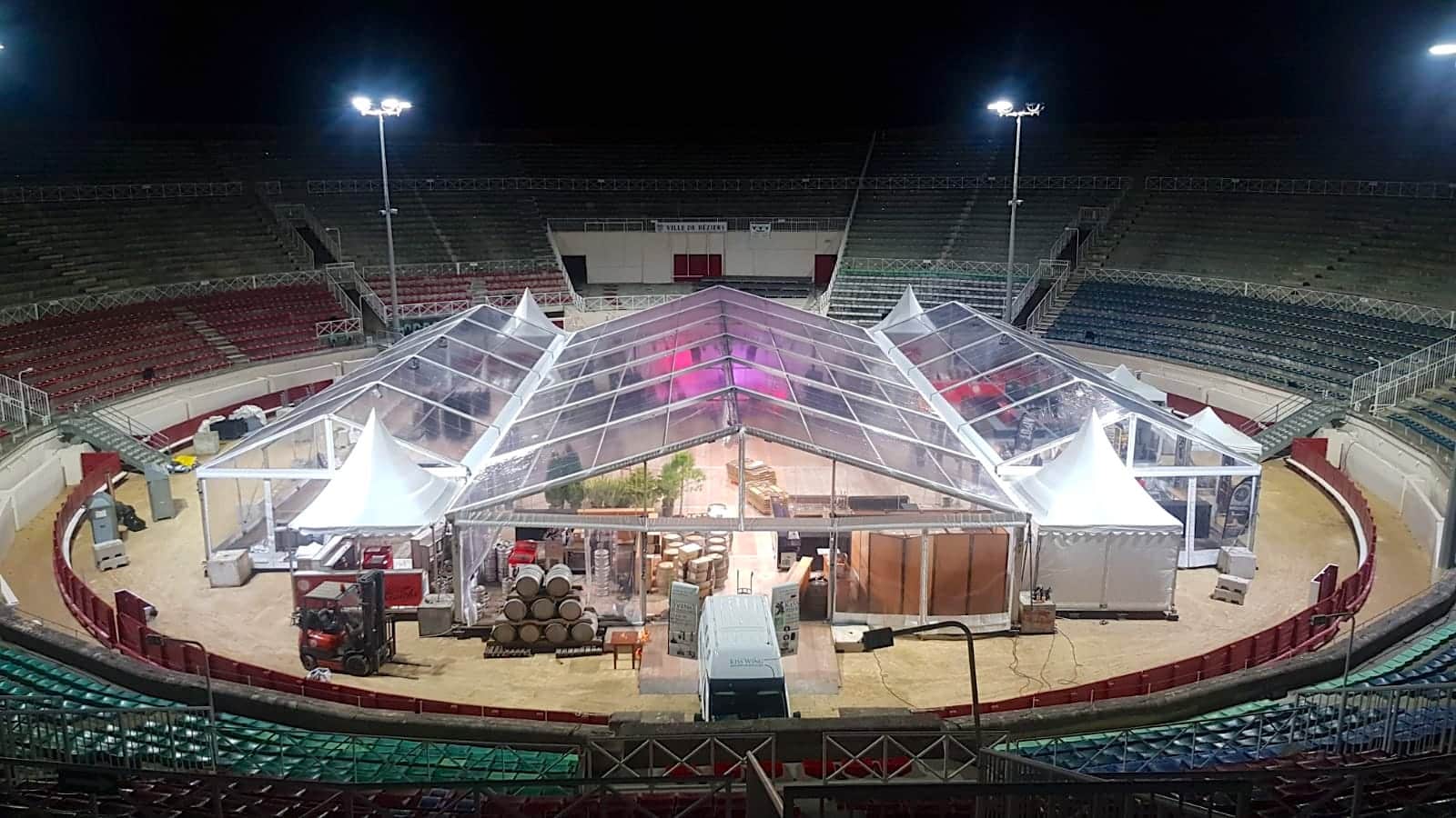 Installation d'une grande tente transparente dans une arène de nuit, vue d'en haut, avec futs de vin et chariots élévateurs. Texte visible: VILLE DE BEZIERS.