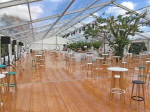 Grande tente événementielle avec toit transparent, plancher en bois lustré, et nombreux mange-debout avec tabourets colorés autour d'un arbre central.