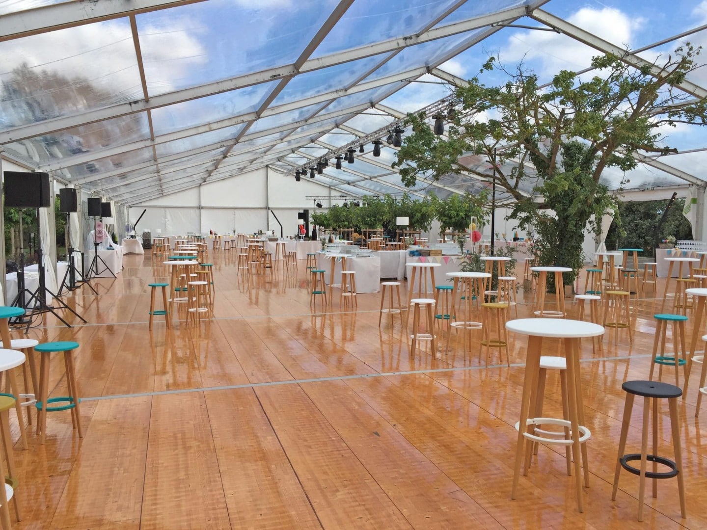 Grande tente événementielle avec toit transparent, plancher en bois lustré, et nombreux mange-debout avec tabourets colorés autour d'un arbre central.