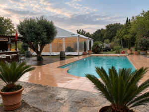 Piscine avec tente de réception éclairée, bar et olivier dans un jardin élégant lors d'une soirée au crépuscule.
