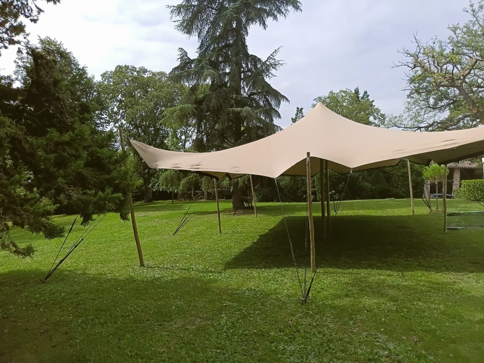 Grande tente stretch beige installée sur une pelouse verte, soutenue par des poteaux en bois, au milieu d'un parc arboré.