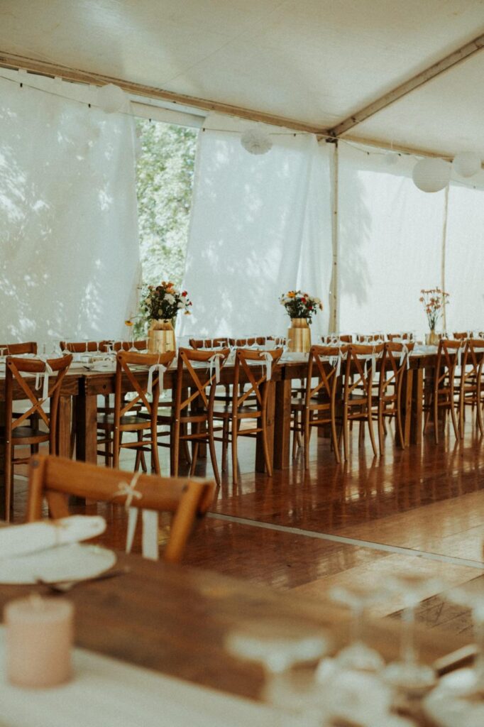Longues tables en bois avec chaises rustiques et décor floral sous un chapiteau clair. Lumière naturelle et ambiance de réception élégante.