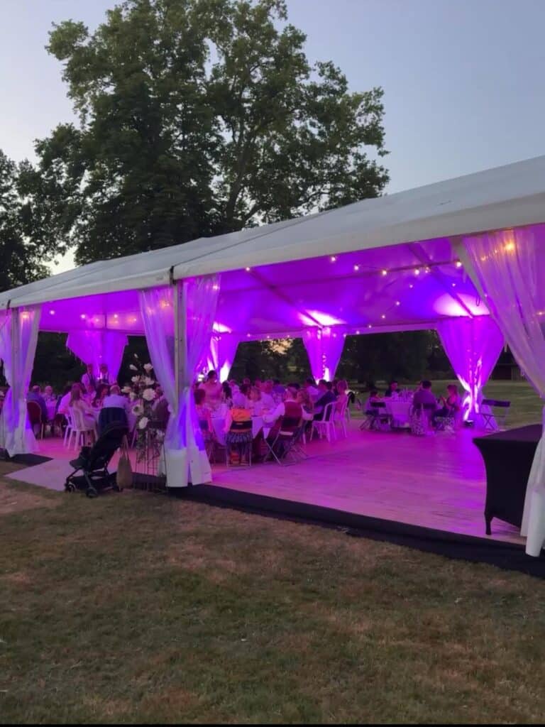 Réception en soirée sous chapiteau blanc éclairé par une lumière violette vive, invités attablés sur plancher en bois.
