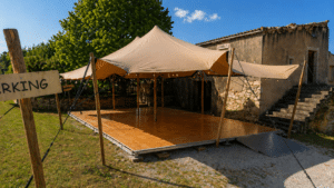 Tente stretch beige sur plancher bois avec guirlandes, panneau PARKING et bâtiment rustique sous ciel bleu.