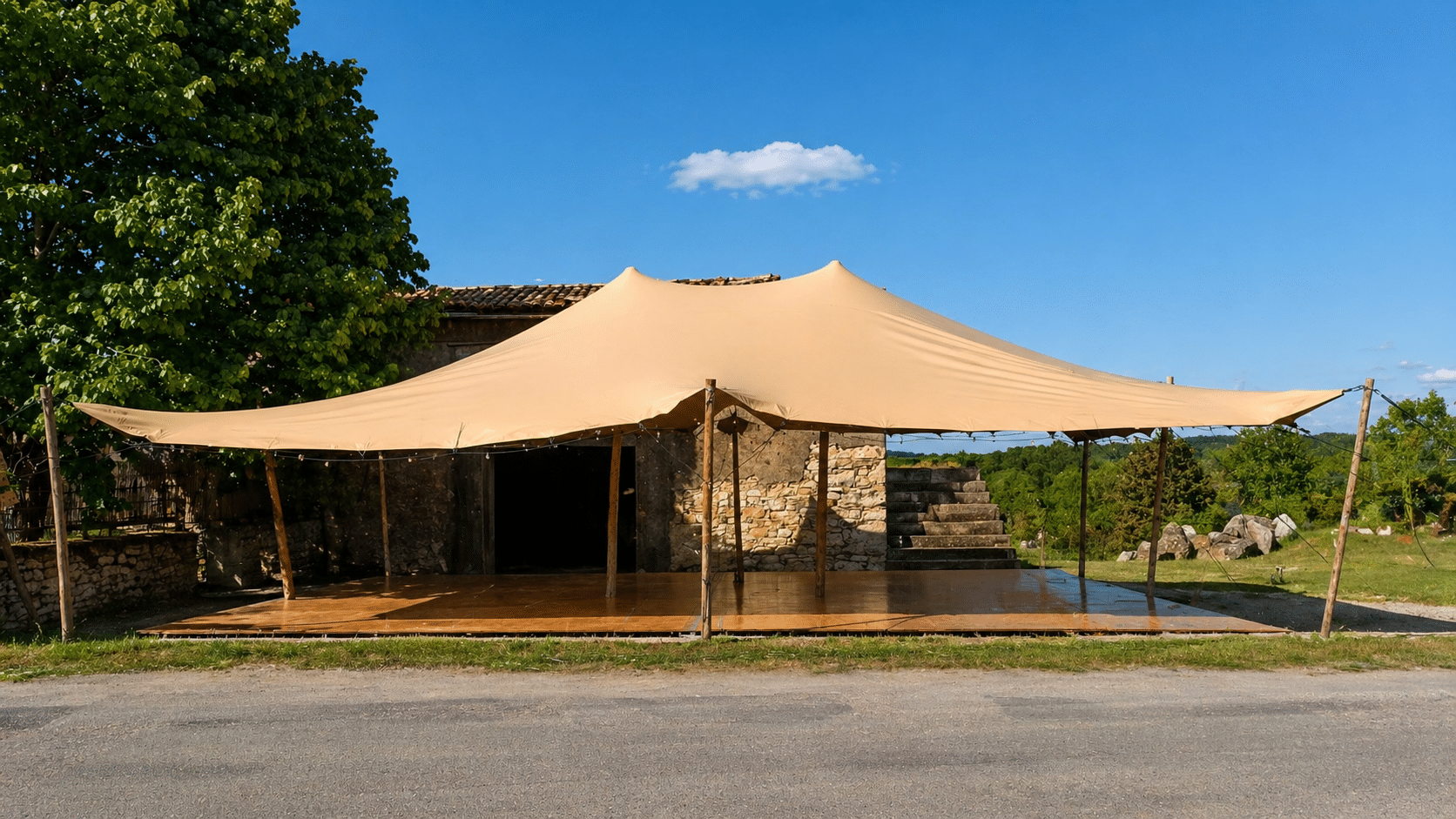 Grande tente stretch beige avec plancher en bois installée devant une bâtisse en pierre sous un ciel bleu.