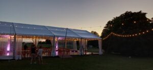 Grand chapiteau blanc illuminé de guirlandes pour un événement en extérieur à la tombée de la nuit.