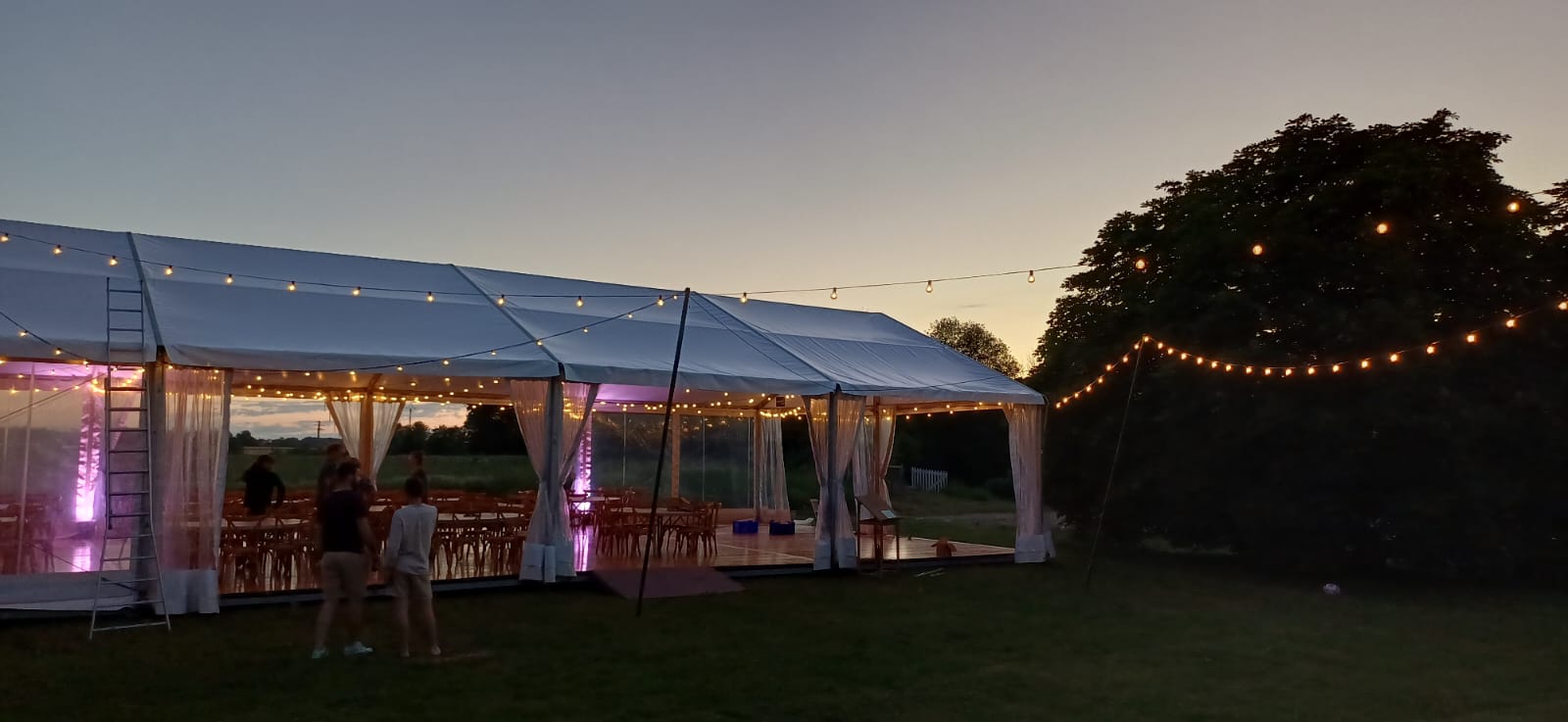 Grand chapiteau blanc illuminé de guirlandes pour un événement en extérieur à la tombée de la nuit.