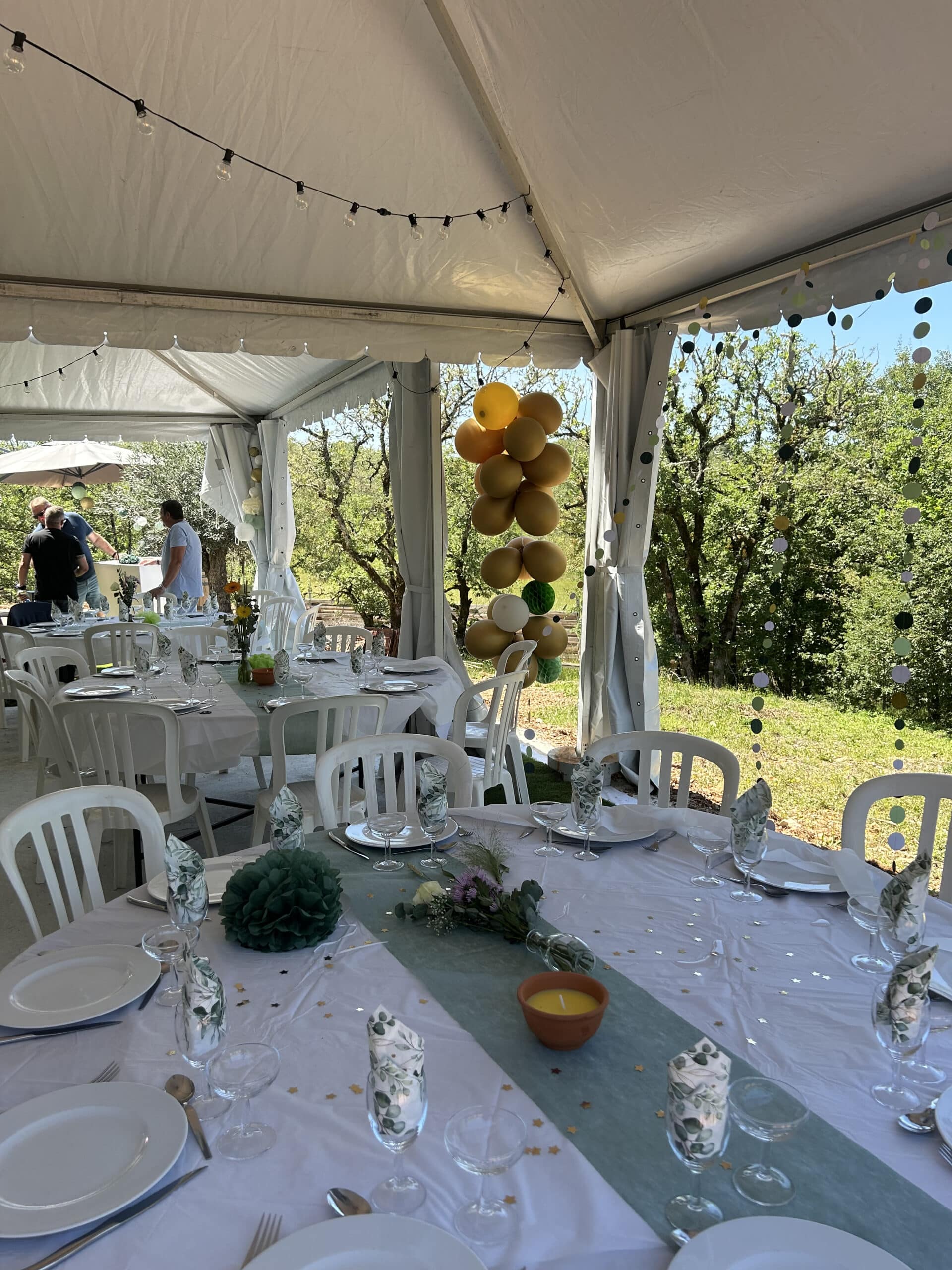 Table de fête décorée sous un chapiteau blanc avec chemin de fer vert sauge, ballons dorés et verres élégants.
