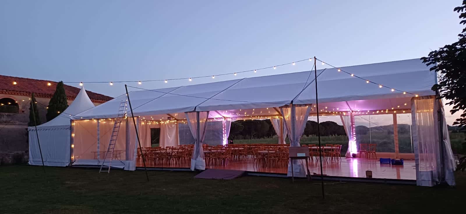 Grande tente de réception blanche illuminée pour un mariage au crépuscule, avec guirlandes et éclairage d'ambiance violet.