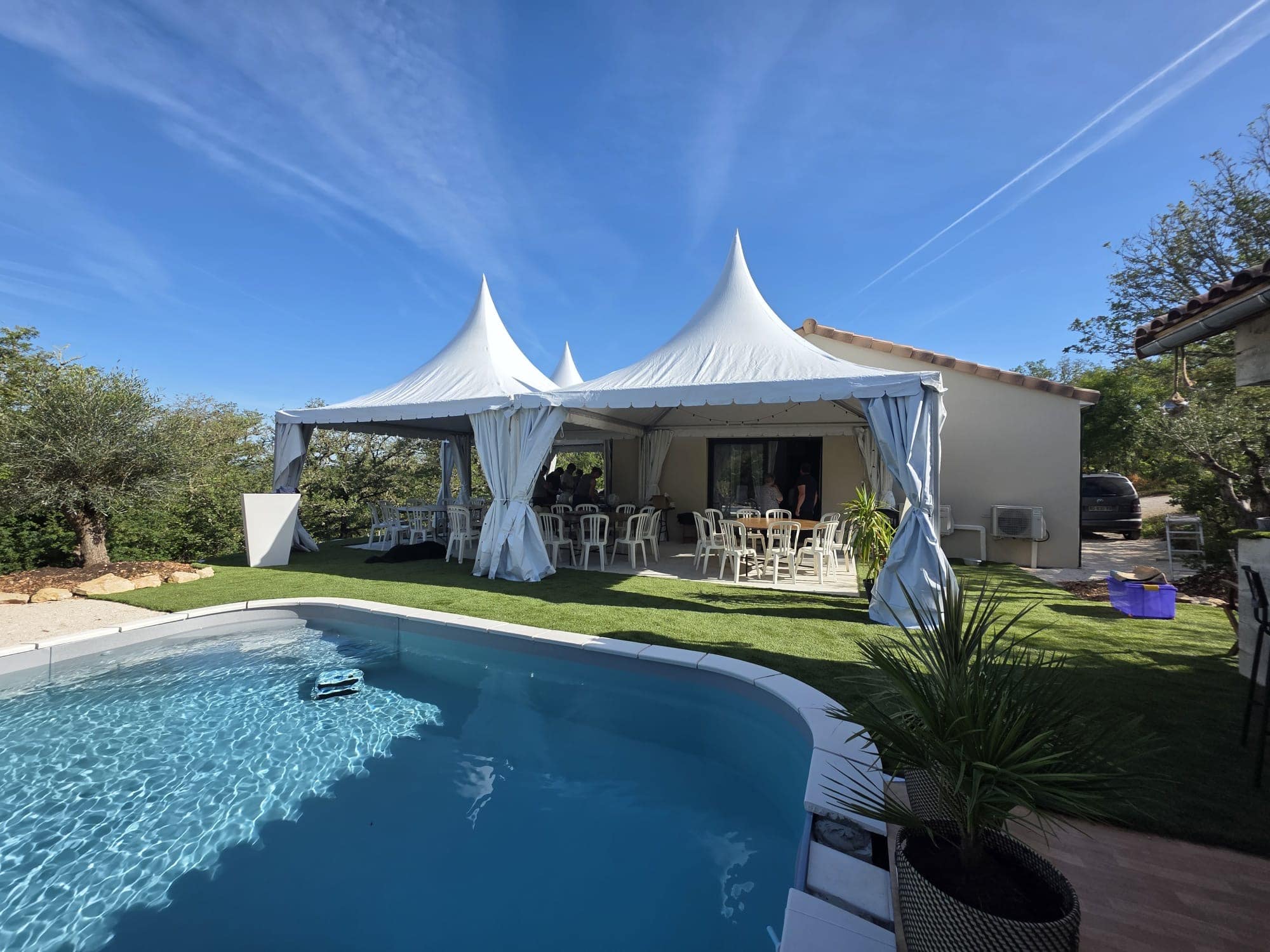 Tentes pagodes blanches pour une réception au bord d'une piscine sur gazon synthétique par grand ciel bleu.