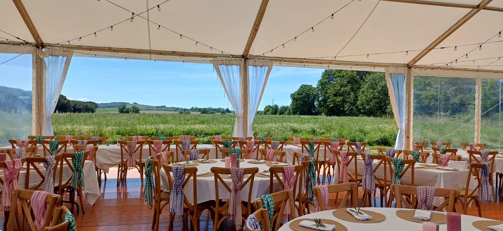 Salle de réception sous chapiteau, tables décorées et chaises en bois avec vue sur une prairie ensoleillée.