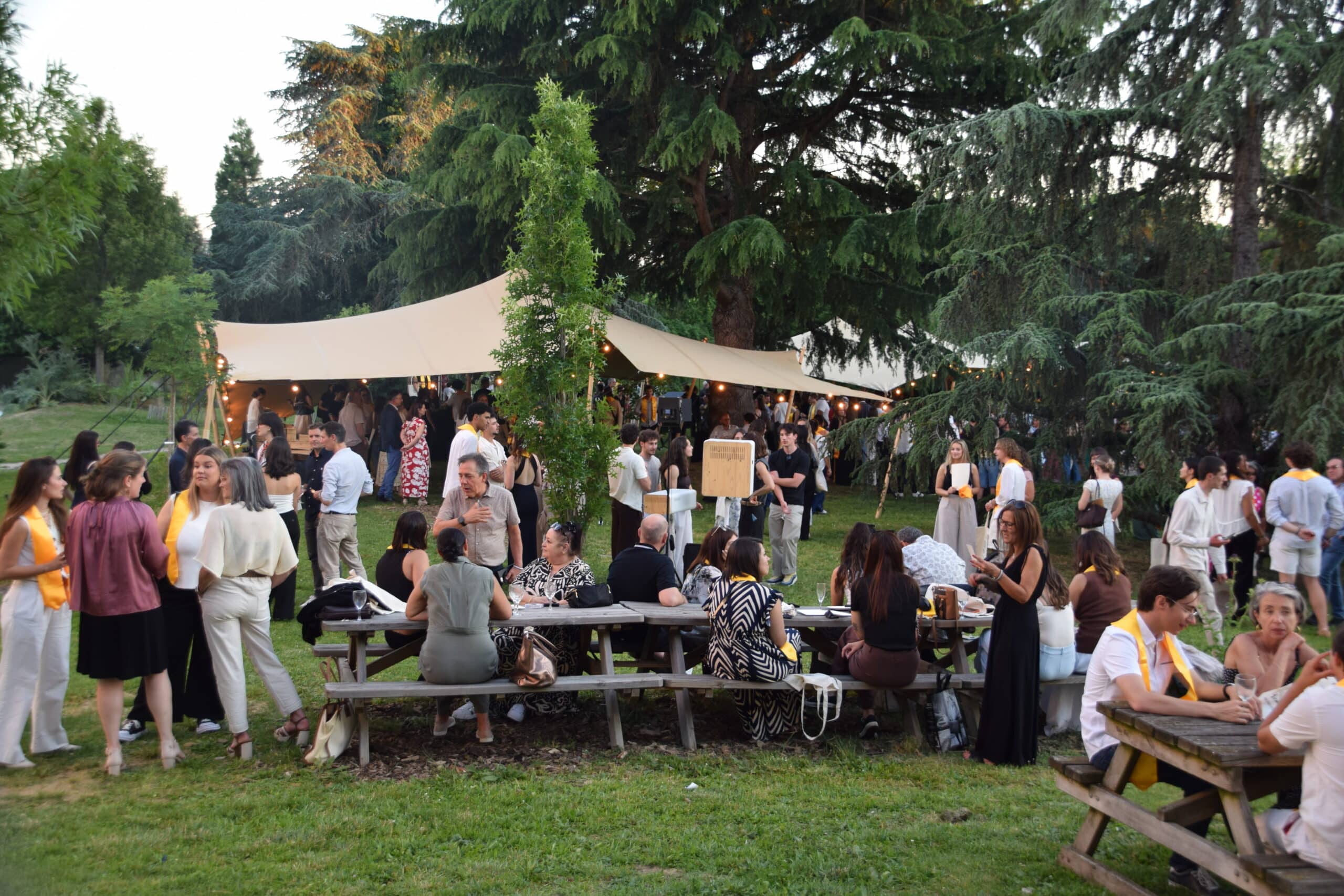 Réception en plein air sous une tente stretch avec des invités en tenue élégante discutant dans un jardin arboré.