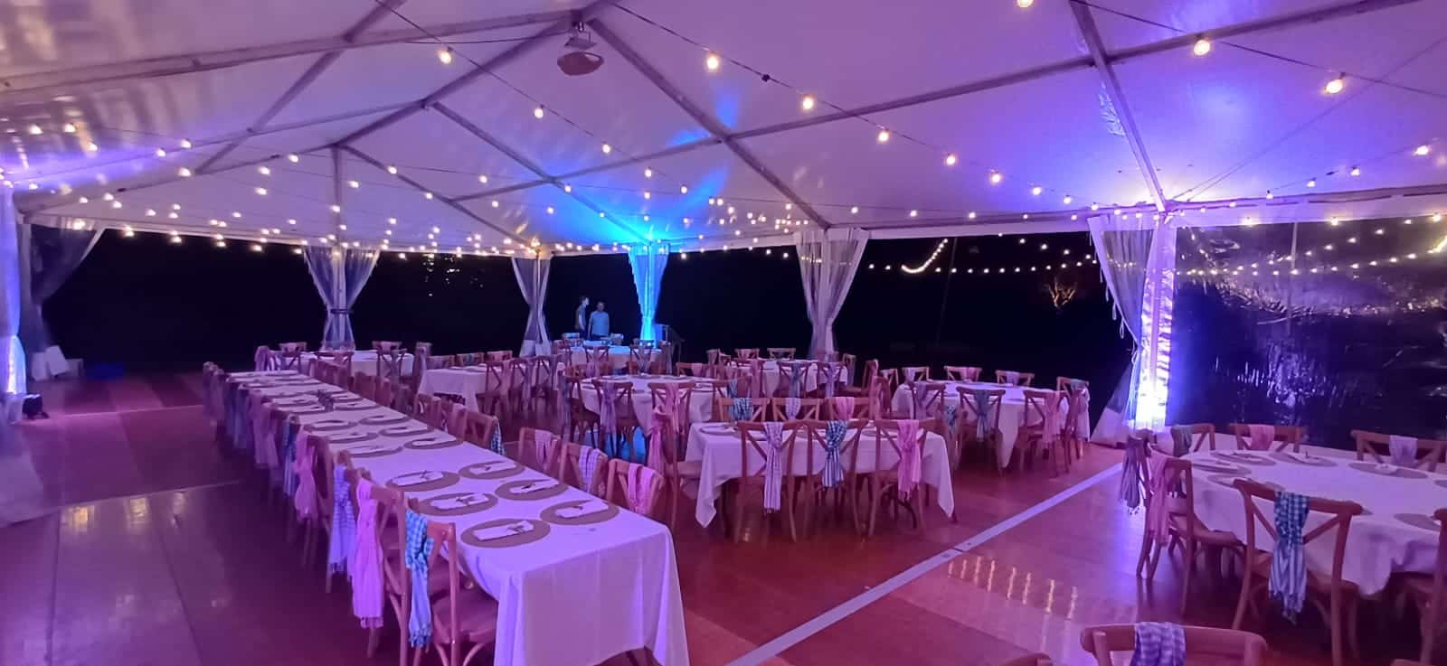 Intérieur de tente de réception illuminée avec tables dressées, chaises décorées et guirlandes lumineuses en soirée.