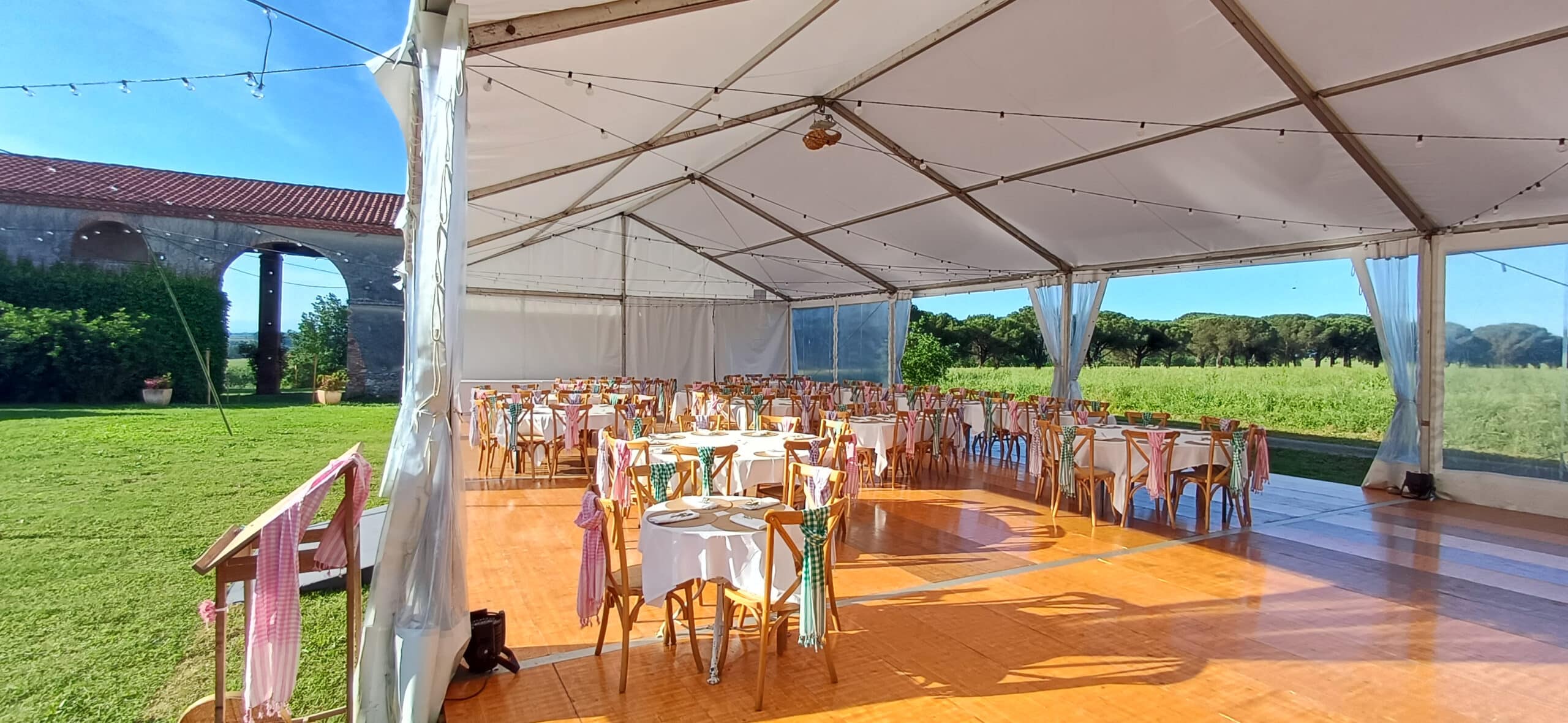 Réception champêtre sous chapiteau blanc avec tables rondes, chaises en bois et foulards colorés en pleine campagne.