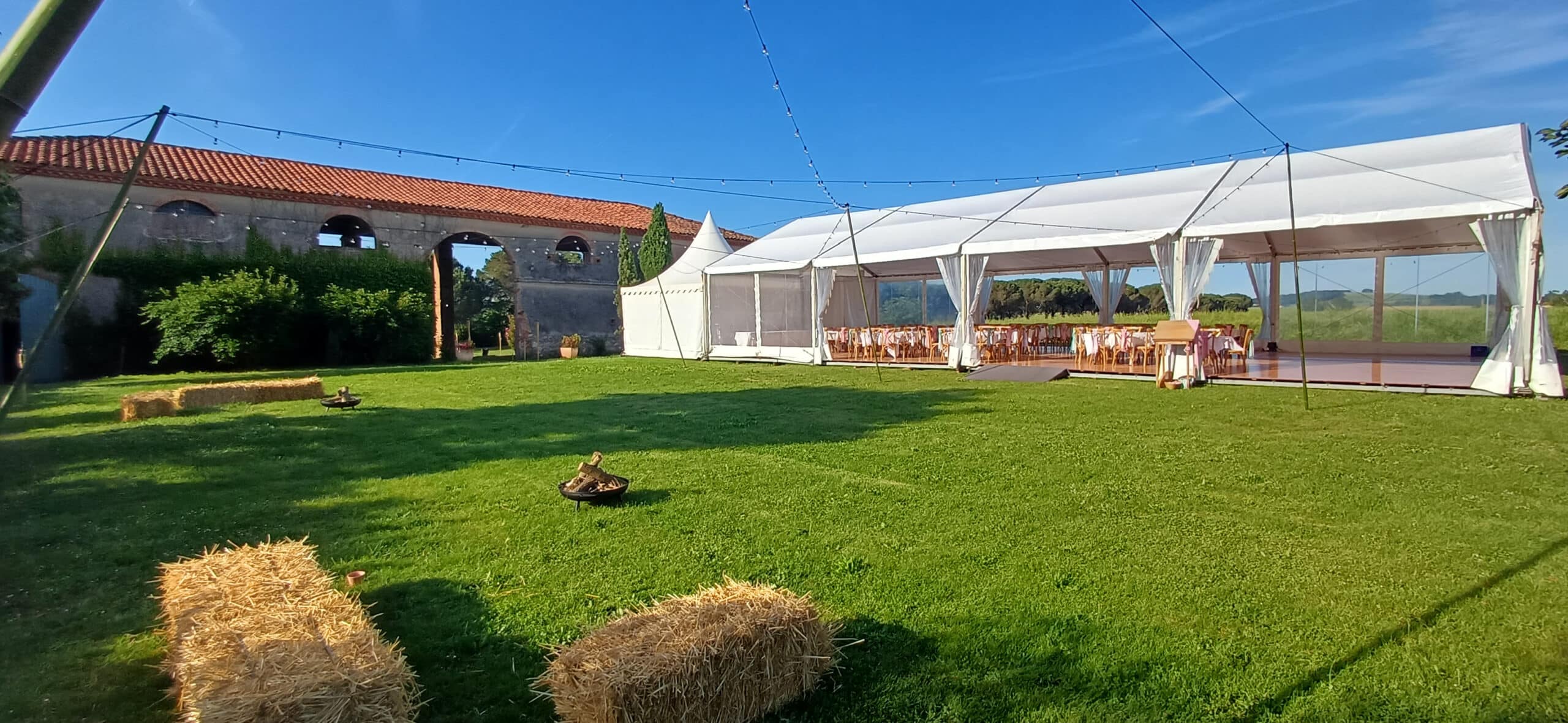 Chapiteau blanc sur pelouse avec bottes de paille et braséro devant un bâtiment ancien sous un ciel bleu dégagé.