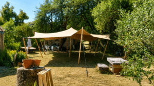 Tente stretch beige dans un jardin arboré avec mobilier et muret en pierre sous le soleil.