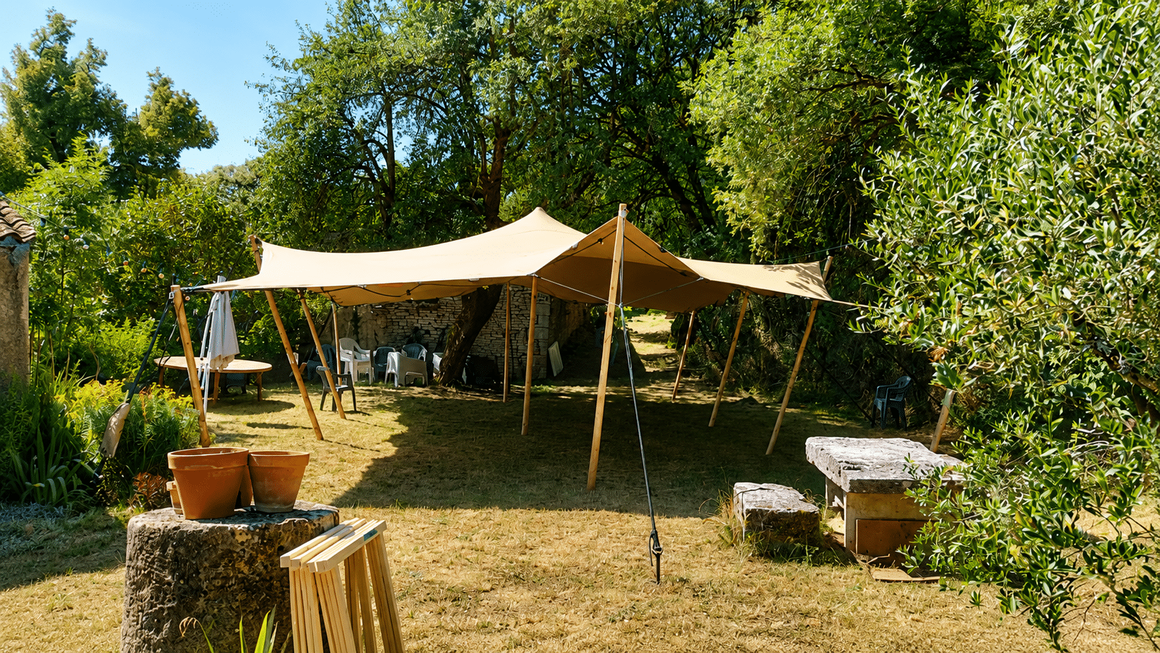 Tente stretch beige dans un jardin arboré avec mobilier et muret en pierre sous le soleil.