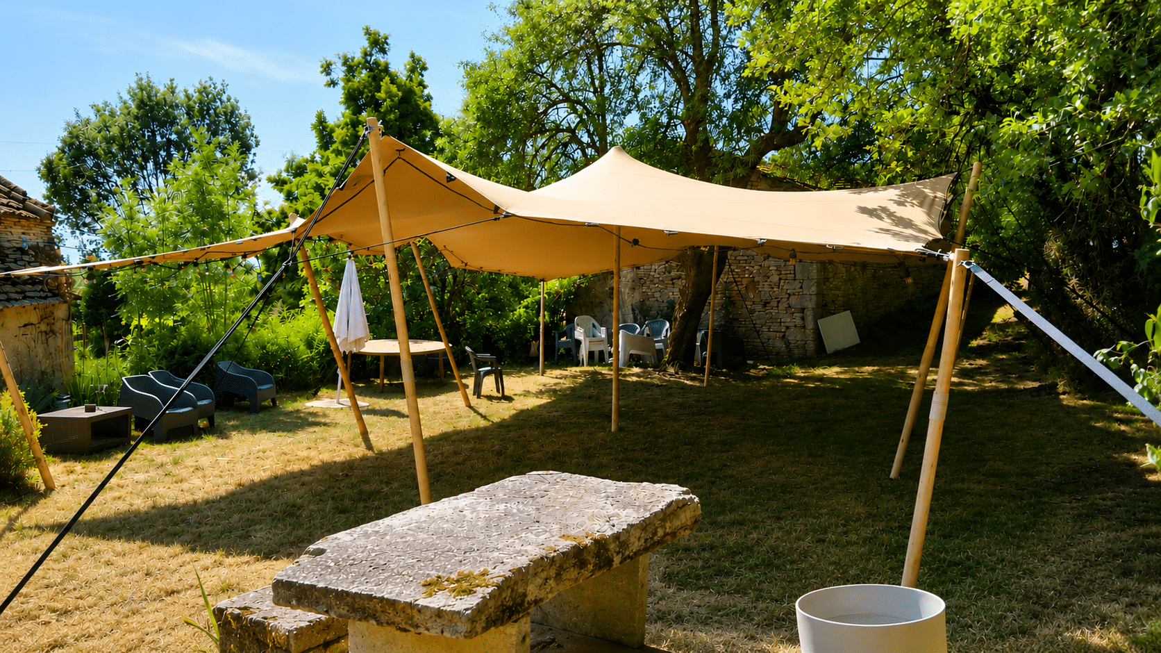 Tente nomade beige dans un jardin ensoleillé avec table en pierre et mobilier extérieur.