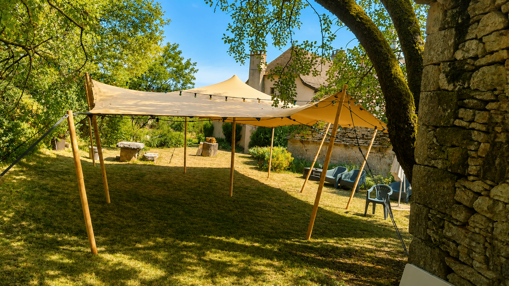 Tente stretch beige dans un jardin ensoleillé avec guirlandes lumineuses et murs en pierre ancienne.