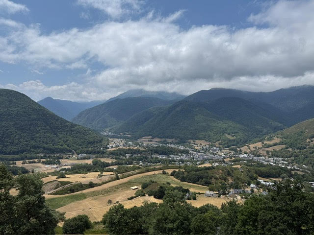 Village niché dans une vallée verdoyante entouré de hautes montagnes sous un ciel nuageux.