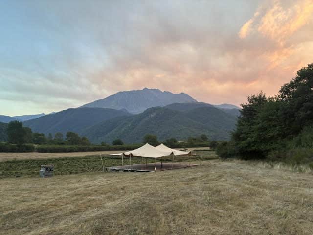 Tente nomade beige sur plateforme en bois face aux montagnes sous un ciel de coucher de soleil rosé.