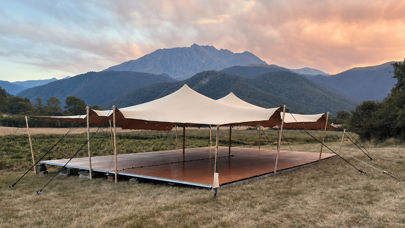 Chapiteau stretch beige et plancher bois face aux montagnes au crépuscule.