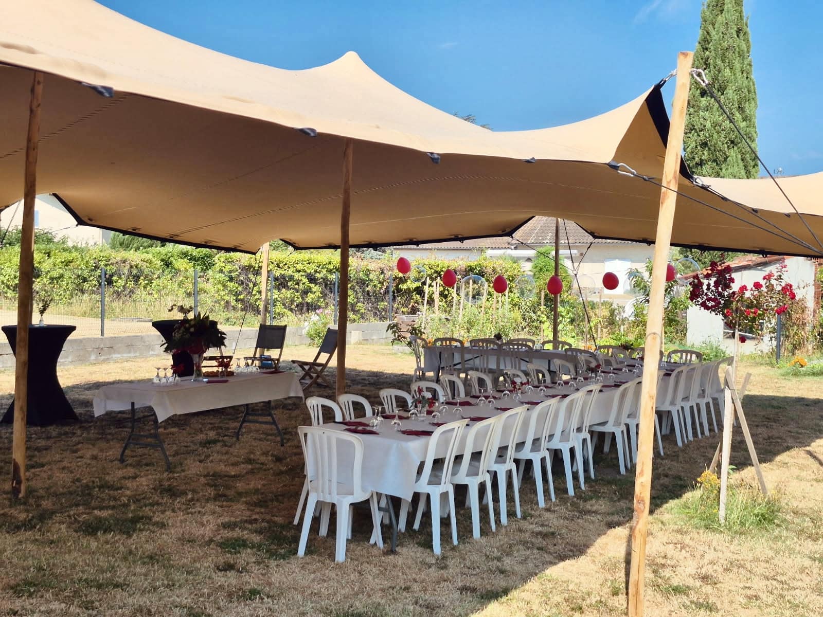 Tente stretch nomade beige, tables de réception nappées et ballons rouges dans un jardin ensoleillé pour un événement.